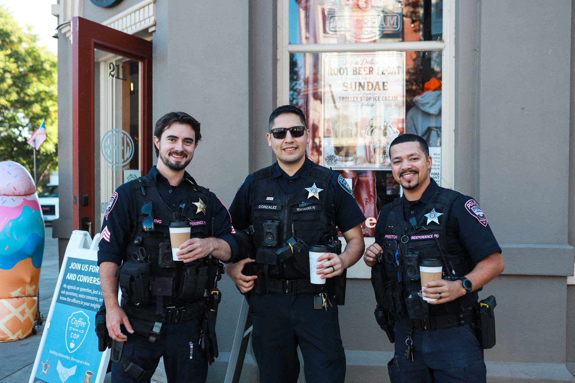 Three smiling police officers in uniform hold coffee cups outside an ice cream shop, posing together in a relaxed, friendly scene.
