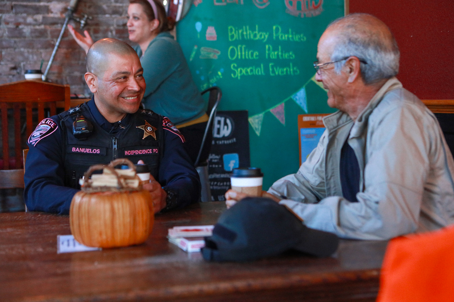 A smiling uniformed officer talks with an elderly man over coffee at a café table; a small pumpkin and event chalkboard create a warm, friendly setting.