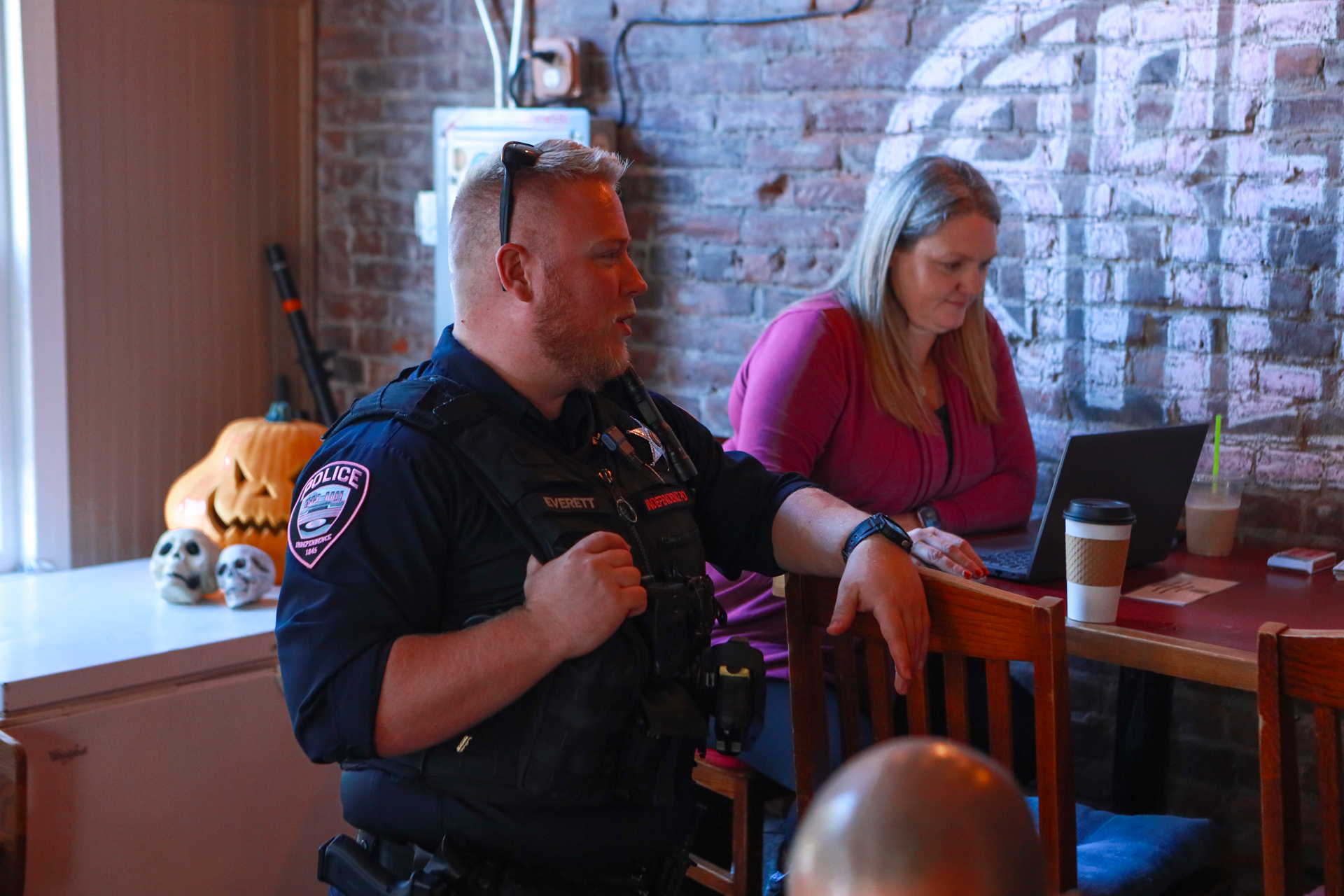 A uniformed police officer gestures while speaking beside a woman using a laptop at a brick-walled table, with Halloween decorations nearby.
