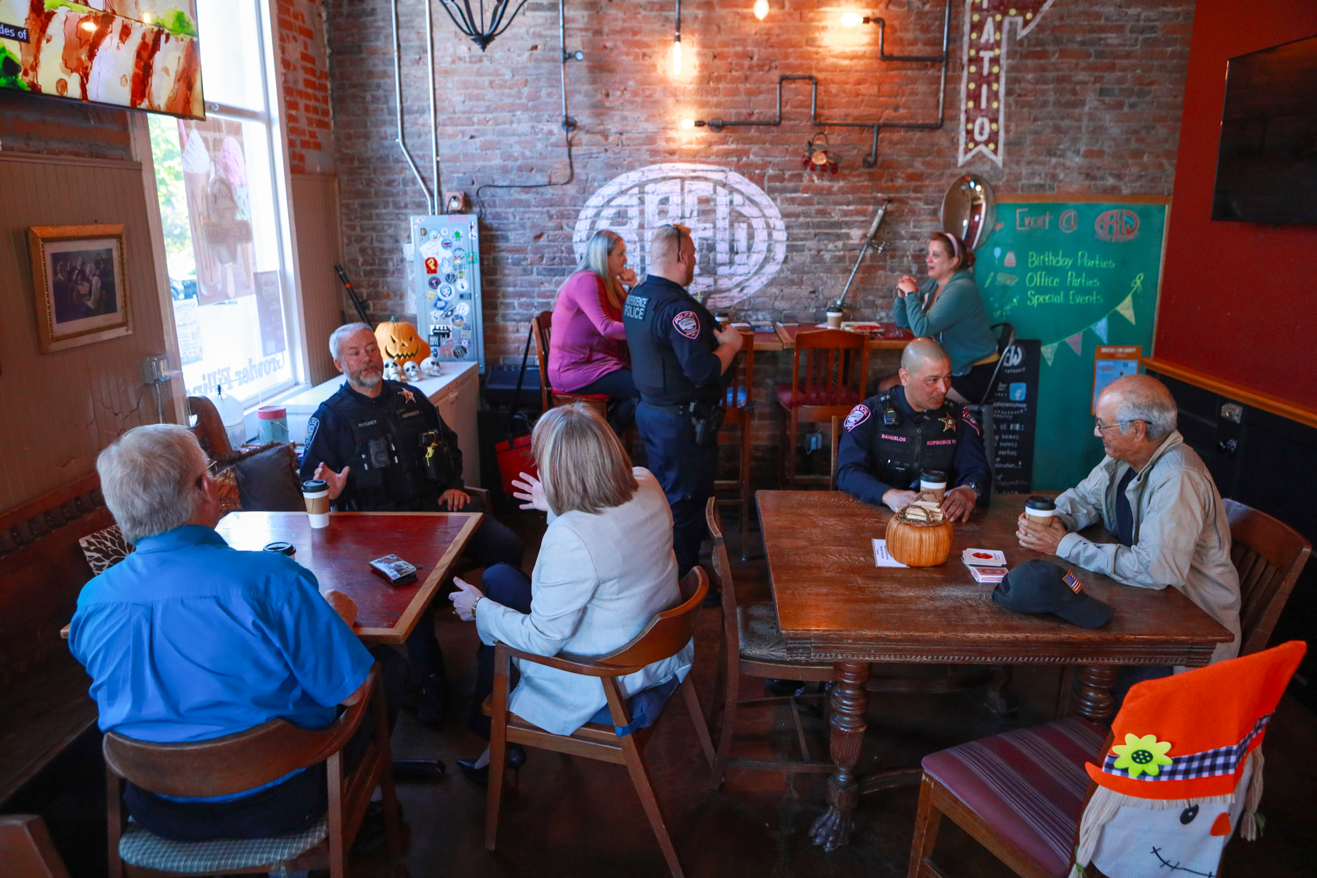 Police officers and community members chat around wooden tables in a warmly lit, brick-walled café, creating a relaxed neighborhood gathering.