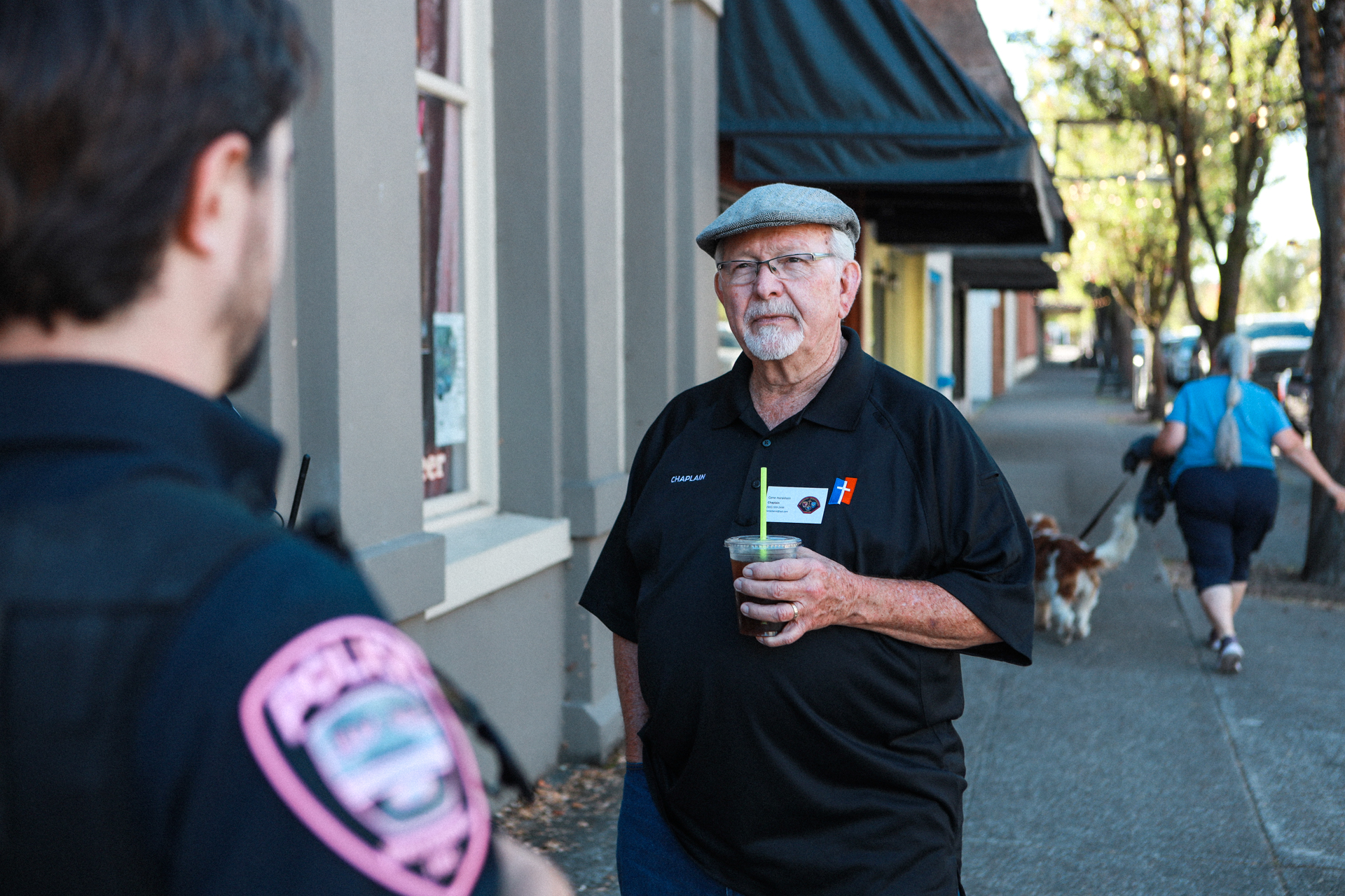 An older man in a flat cap holds a drink and talks with a uniformed officer on a quiet sidewalk; passersby and a dog move behind them.