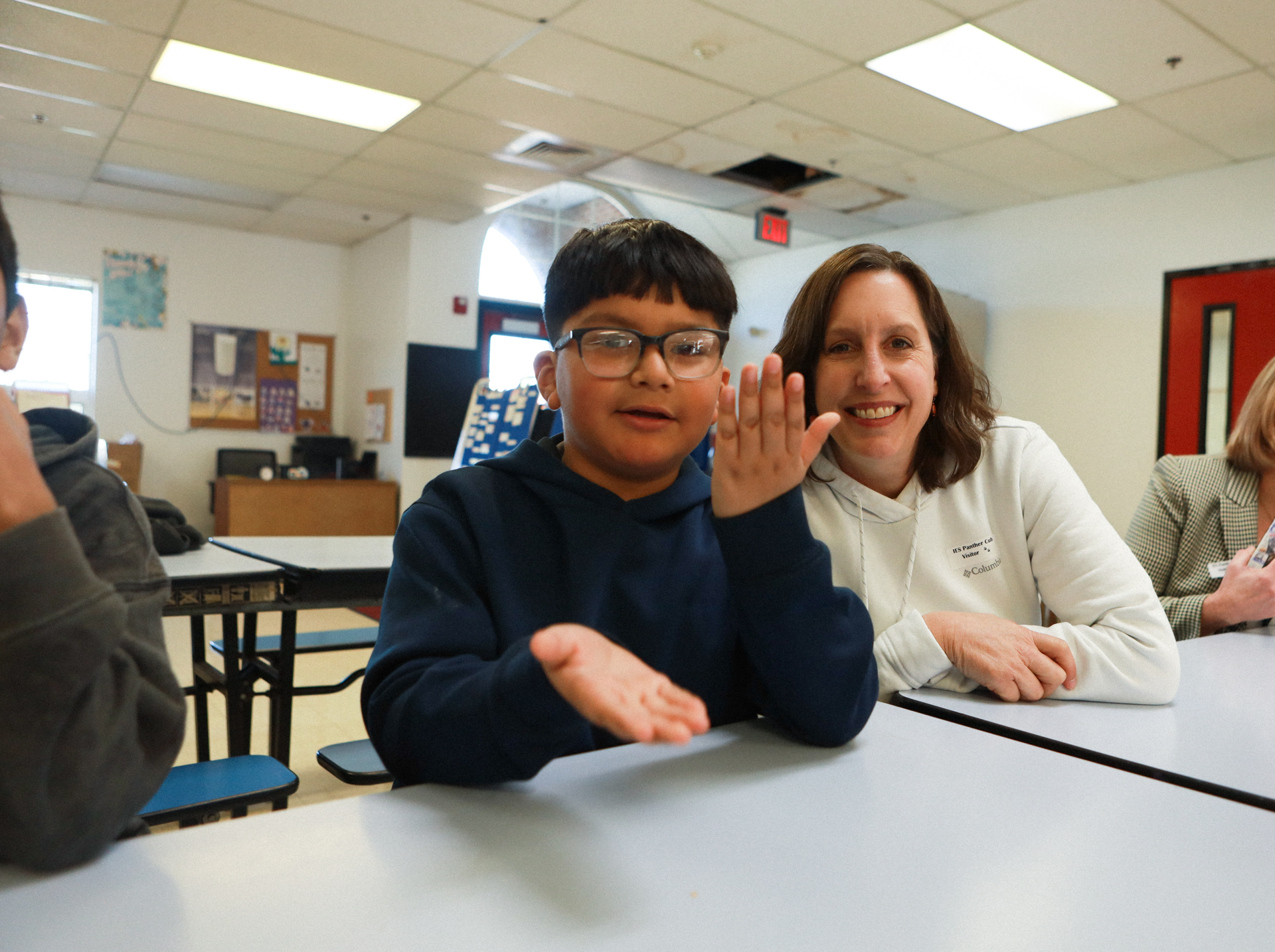 A smiling student in glasses gestures with both hands beside a supportive teacher at a classroom table; their friendly interaction feels warm and welcoming.