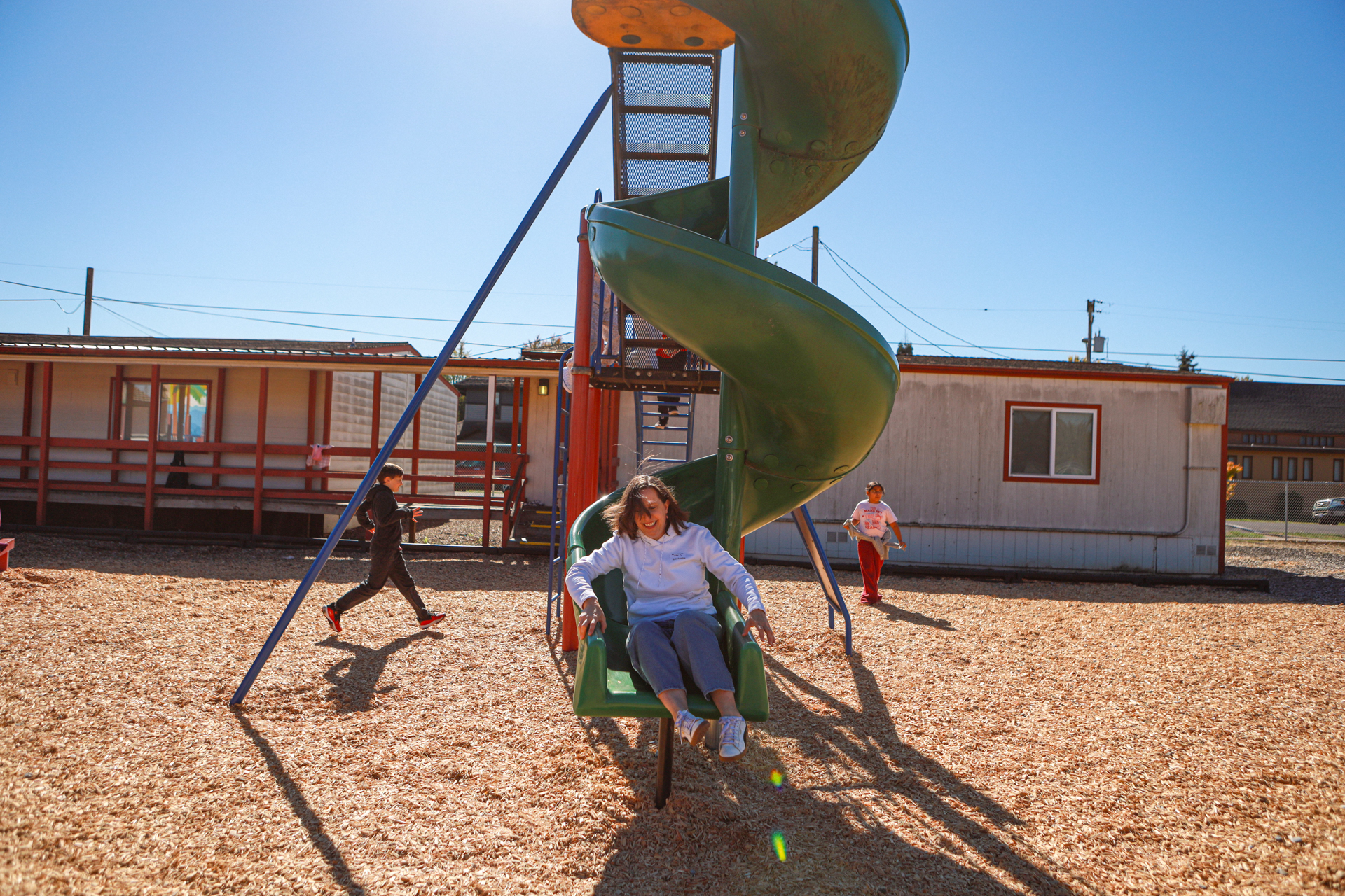 A woman descends a tall green spiral slide as two others play nearby in a sunny schoolyard, casting long shadows and creating a lively, carefree scene.