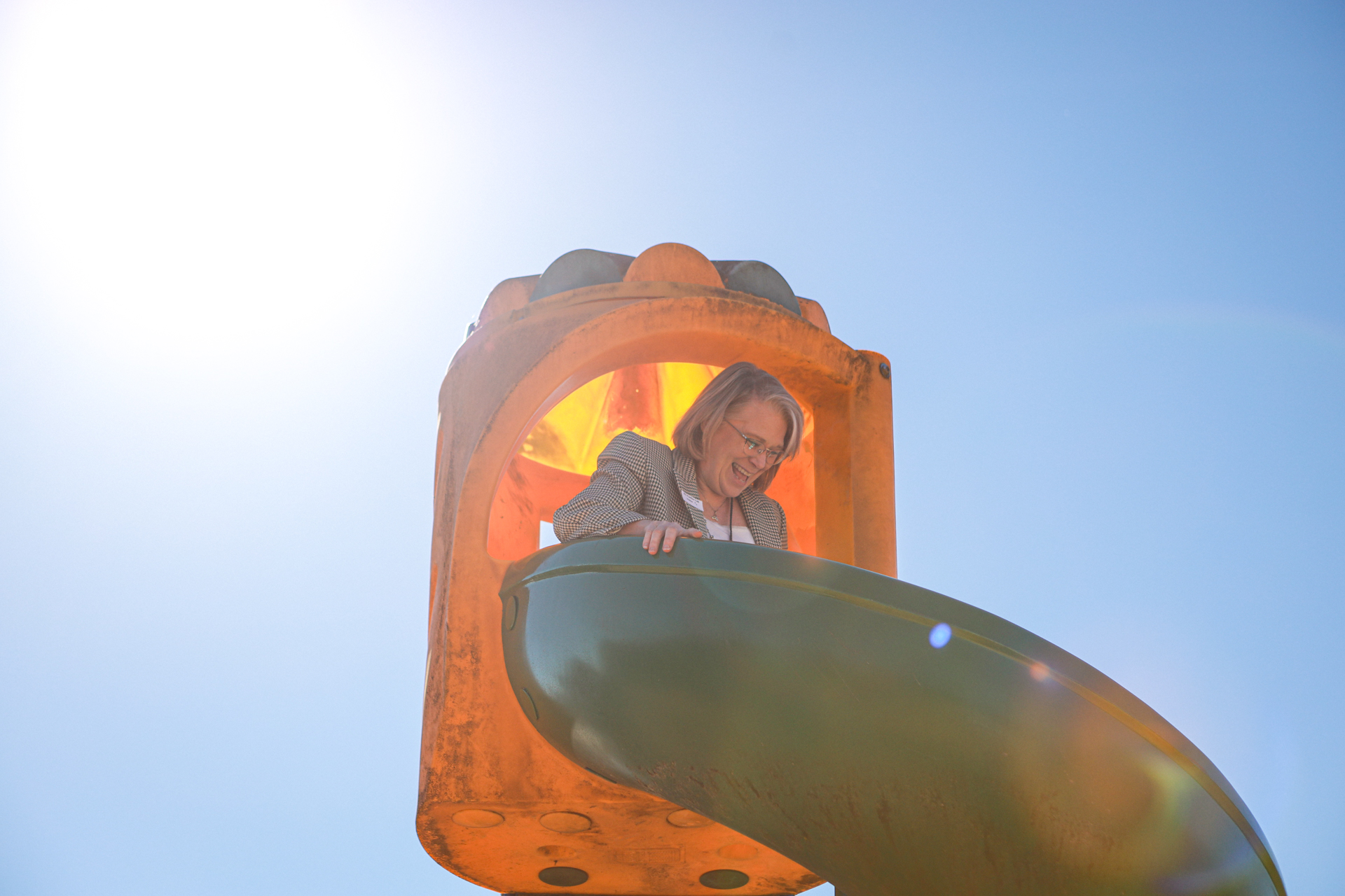 A smiling woman in glasses leans from the top of an orange playground slide, backlit by bright sunlight against a clear blue sky. Playful, warm mood.