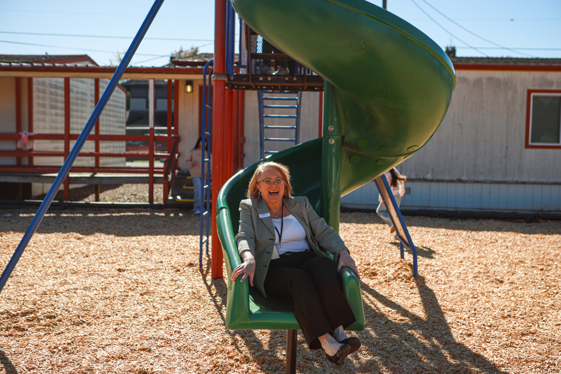 A smiling woman slides down a green playground slide into wood-chip-covered ground, enjoying a sunny day at an outdoor play area.