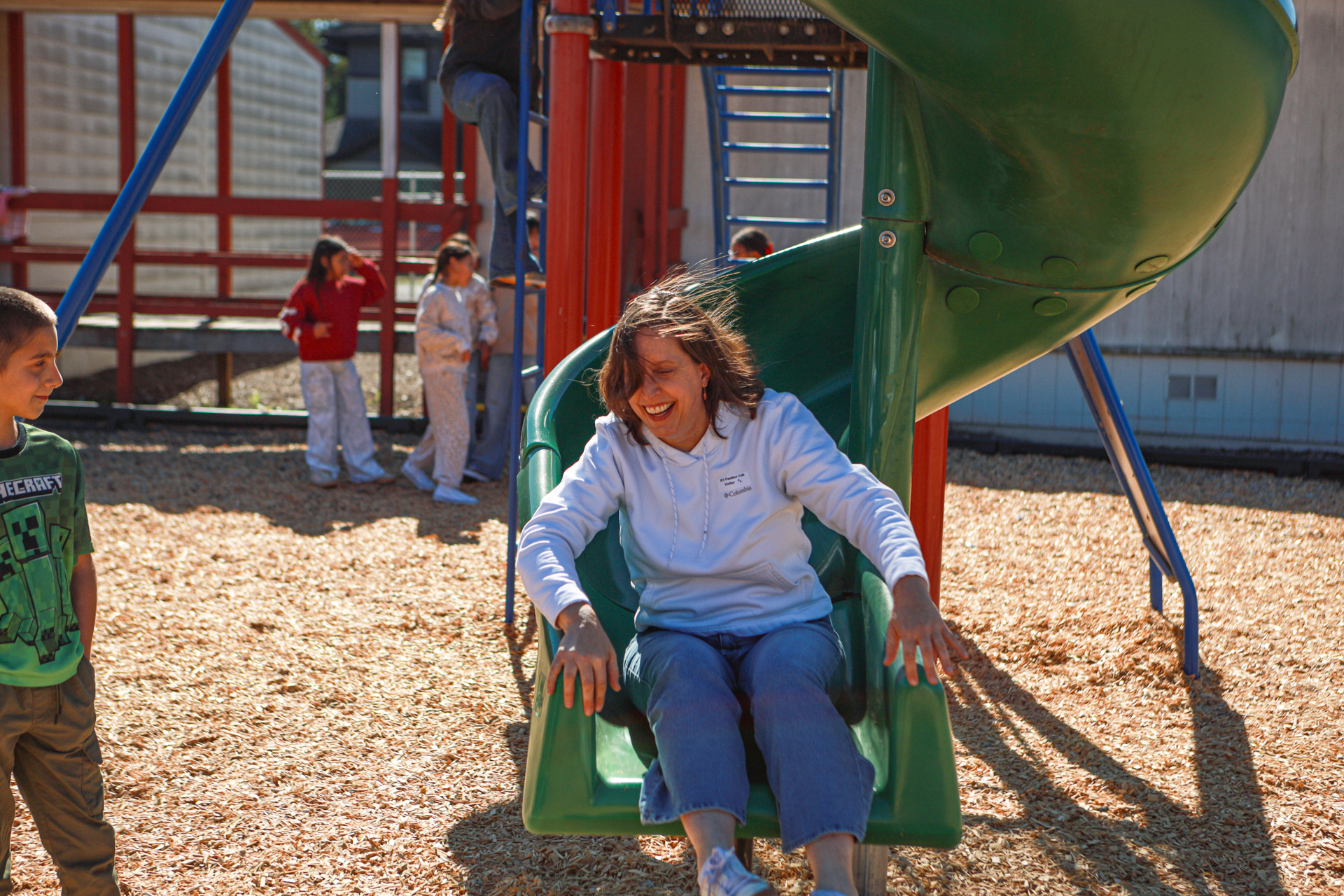 A smiling woman in a white hoodie slides down a green playground slide as children watch nearby, creating a cheerful, playful scene in bright sunlight.