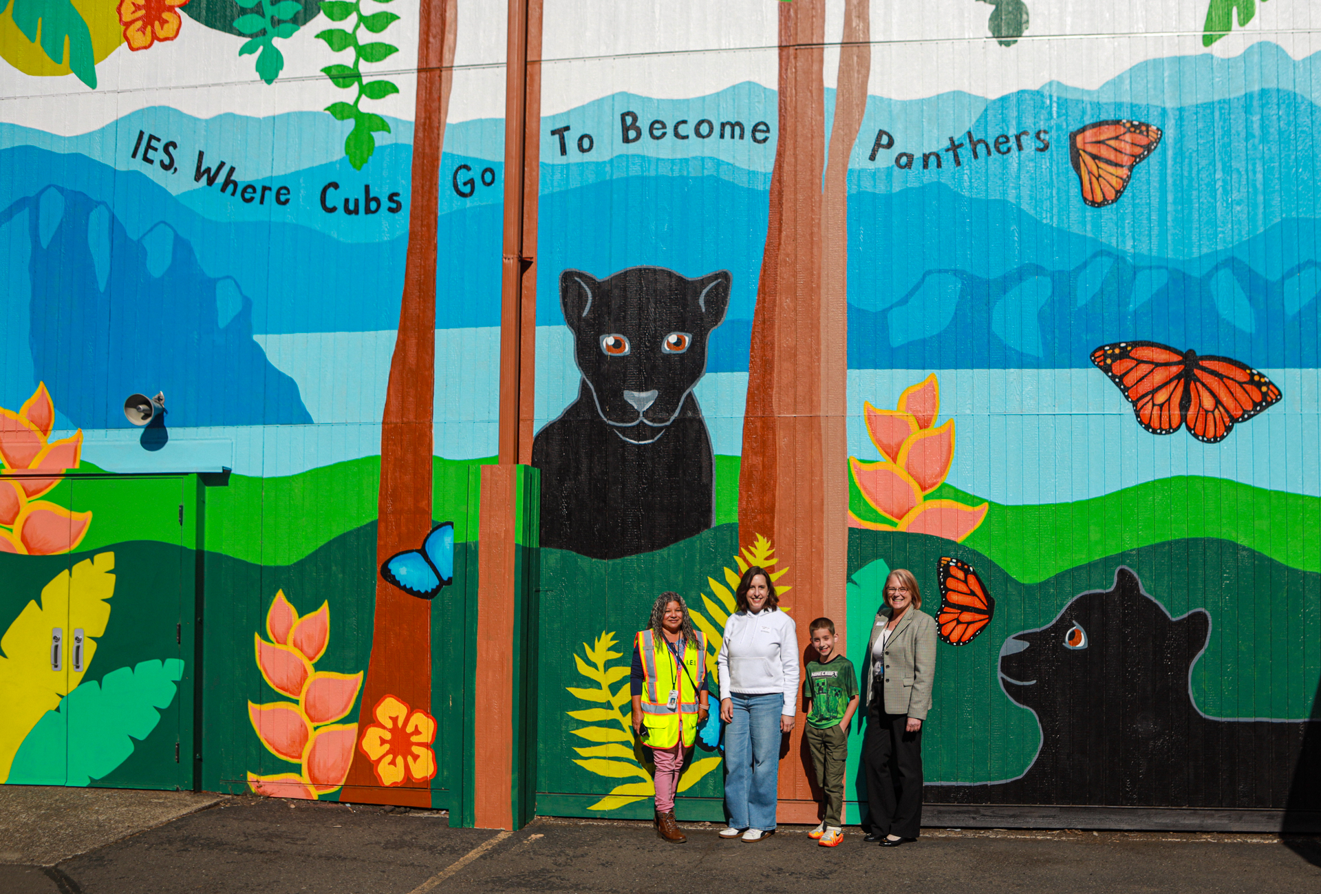 Four people pose before a vibrant school mural showing a black panther, cub, butterflies, flowers, and the words “Yes, Where Cubs Go To Become Panthers.”