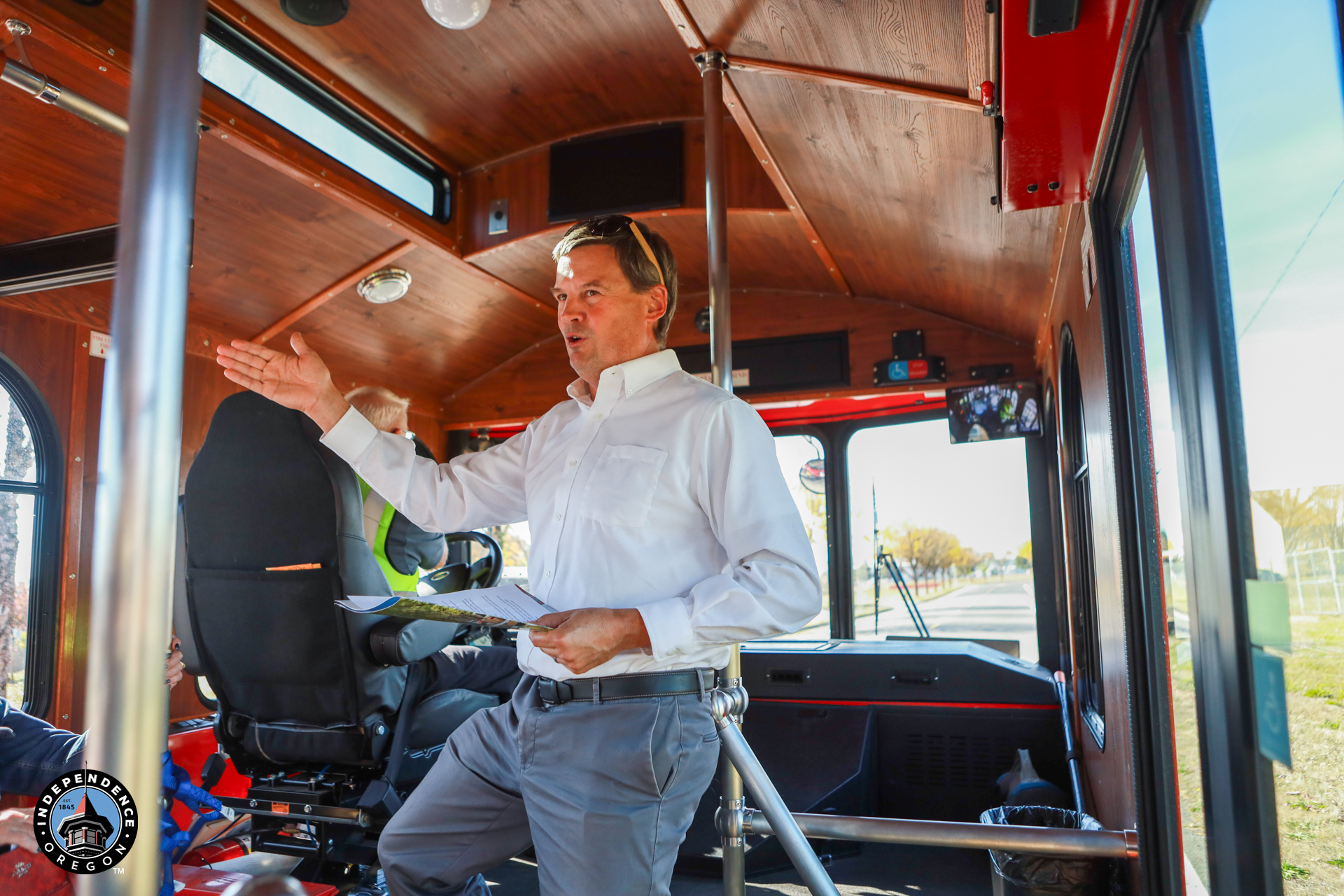A man in a white shirt gestures while explaining the wood-paneled interior of a vintage bus, with driver’s seats and large windows visible.