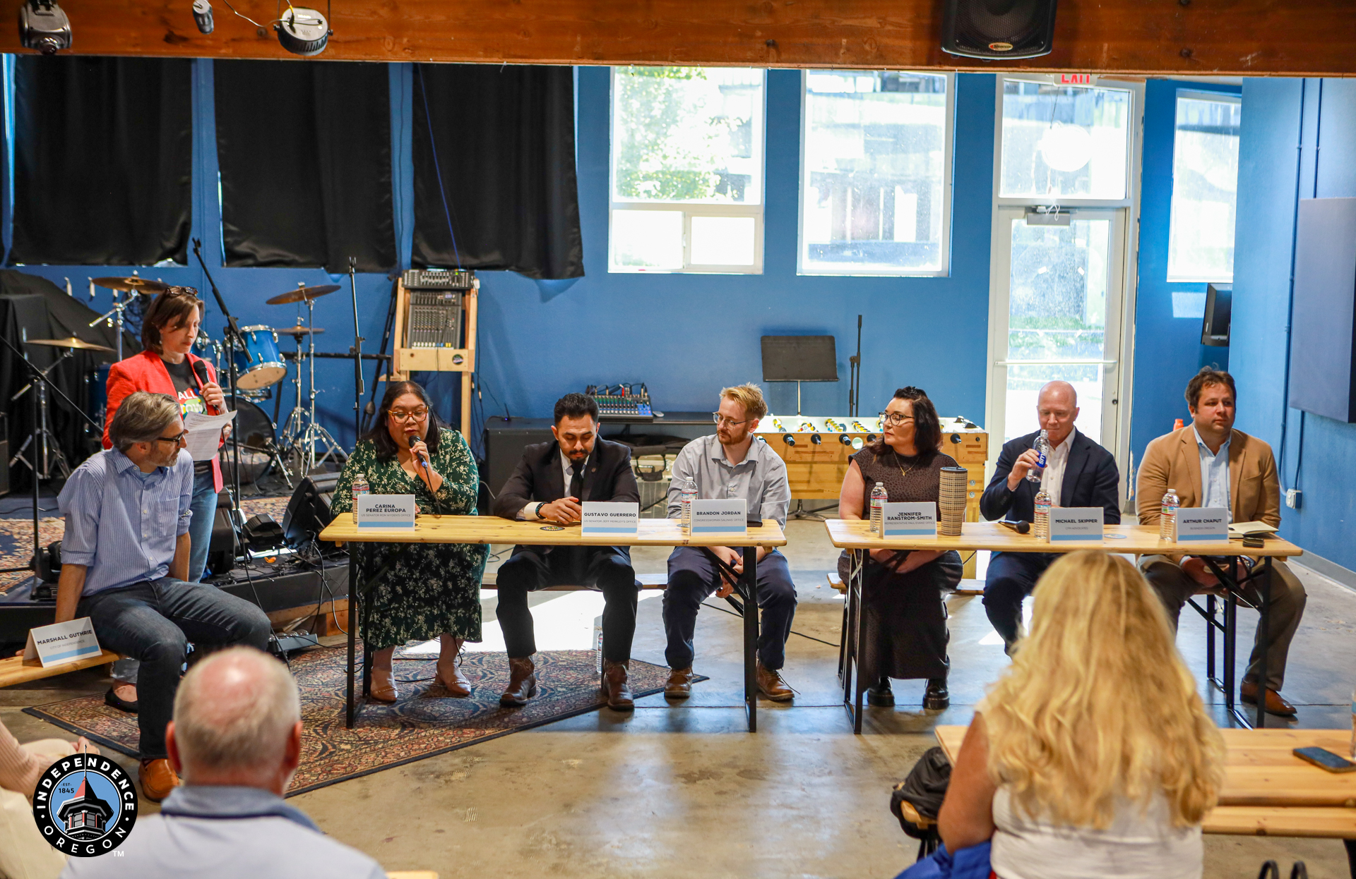 A diverse panel of speakers sits behind a long table in a bright blue room, addressing a small audience during a calm community discussion.