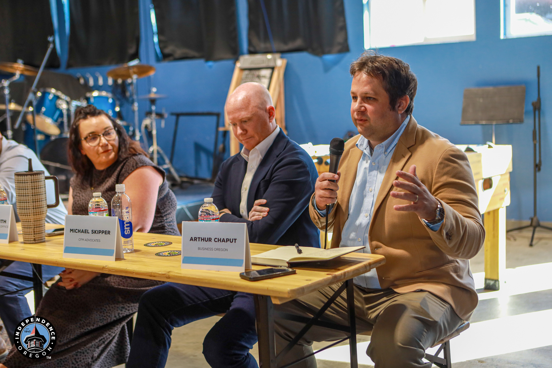 At a studio panel, a man in a tan blazer speaks into a microphone beside two guests, with name placards on the table and a blue backdrop. The mood is attentive.