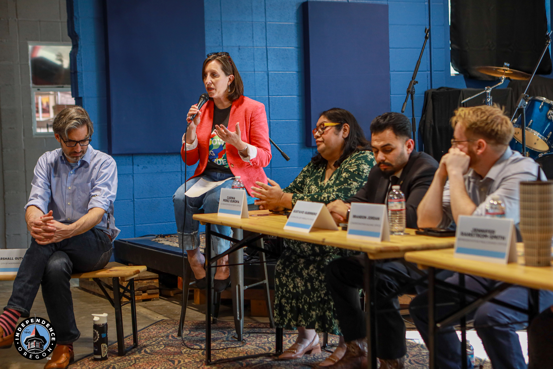 A diverse panel of speakers sits behind a long table in a bright blue room, addressing a small audience during a calm community discussion.