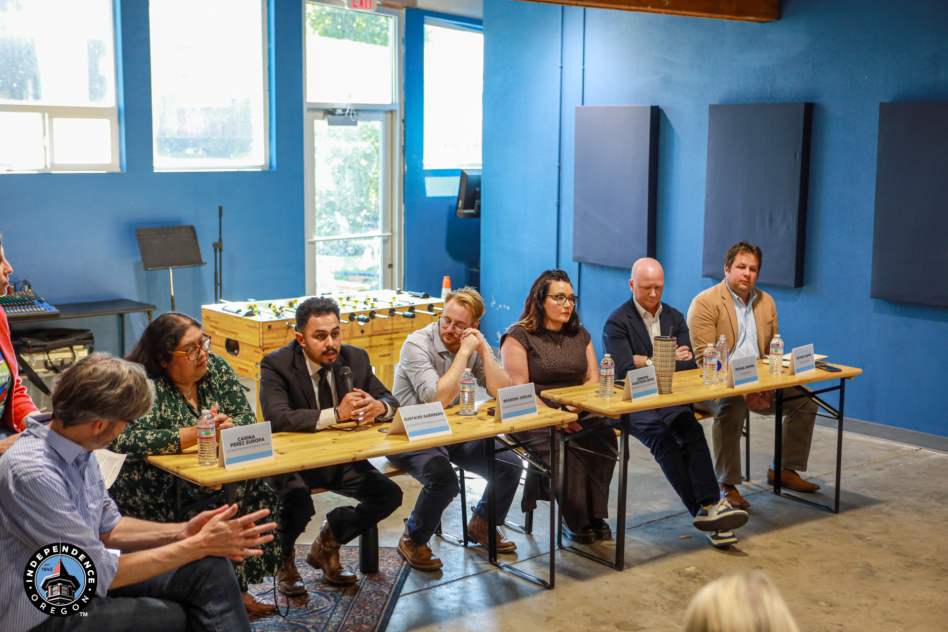 A diverse panel of speakers sits behind a long table in a bright blue room, addressing a small audience during a calm community discussion.