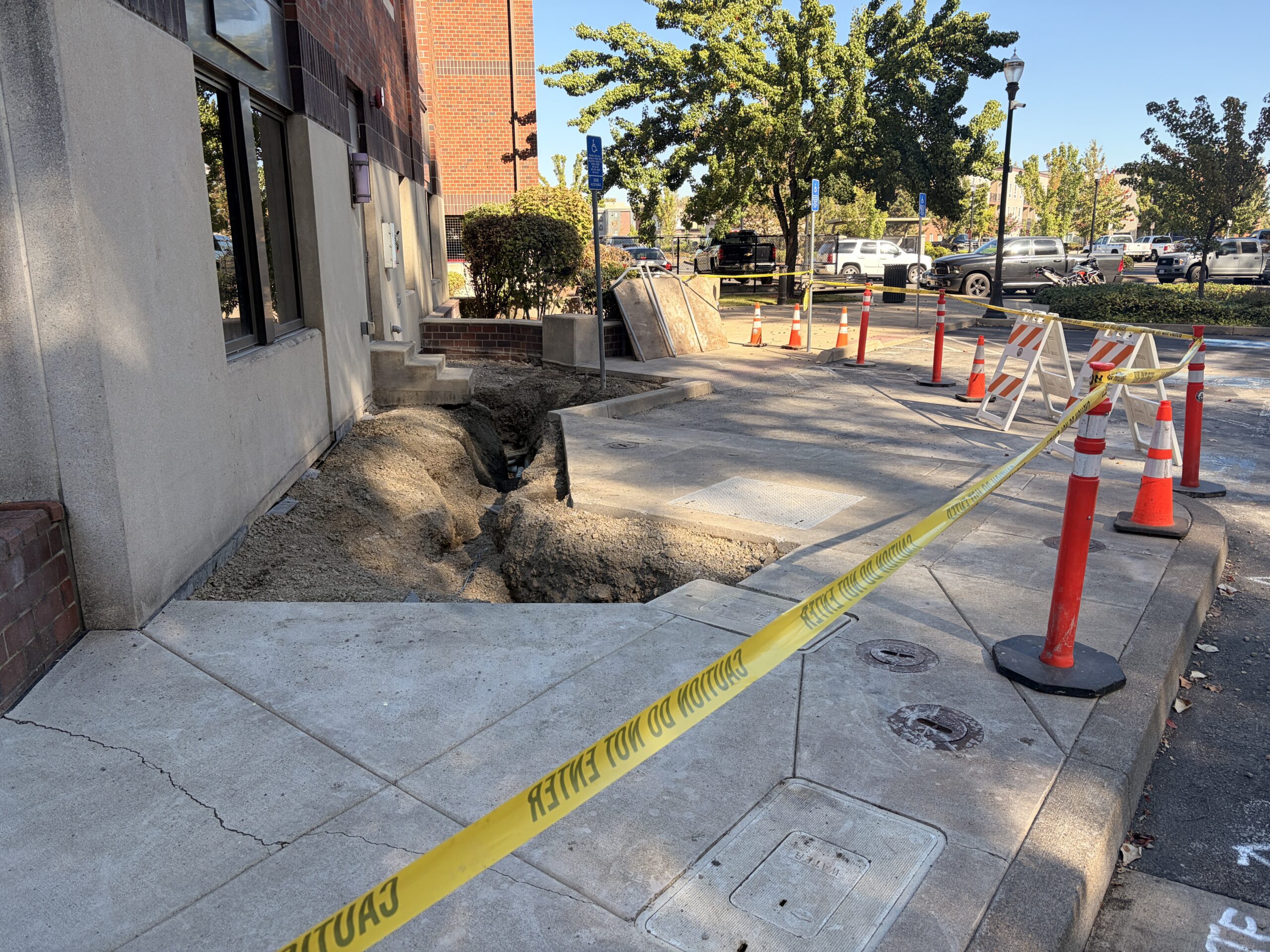 Construction zone beside a brick-and-stucco building, with an excavated sidewalk trench cordoned off by yellow caution tape and orange cones in daylight.